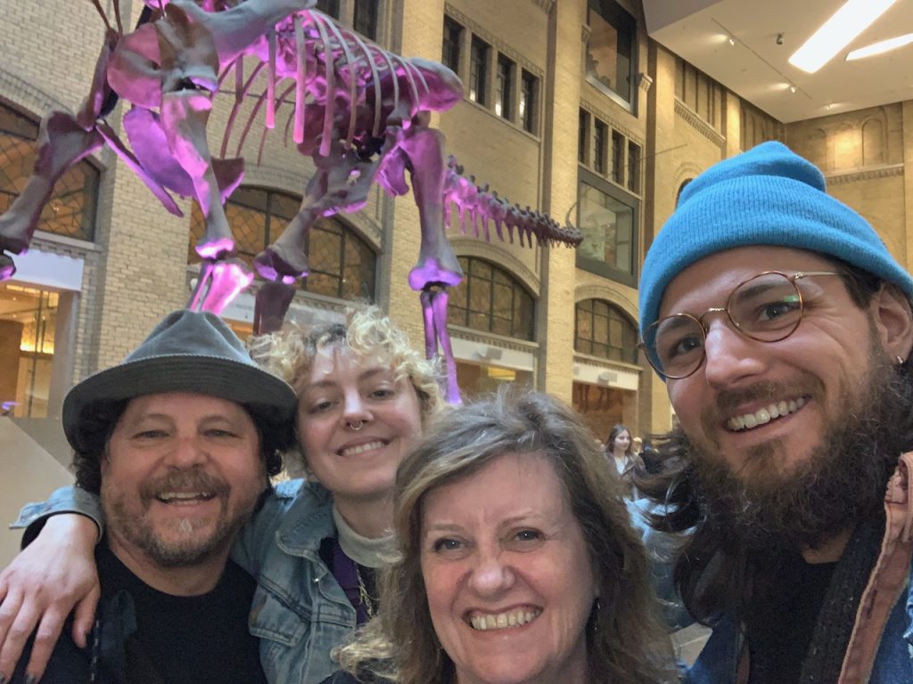 Four people of varying ages, on in a fedora and one in a blue toque smiling at the camera with a purple dinosaur in the background