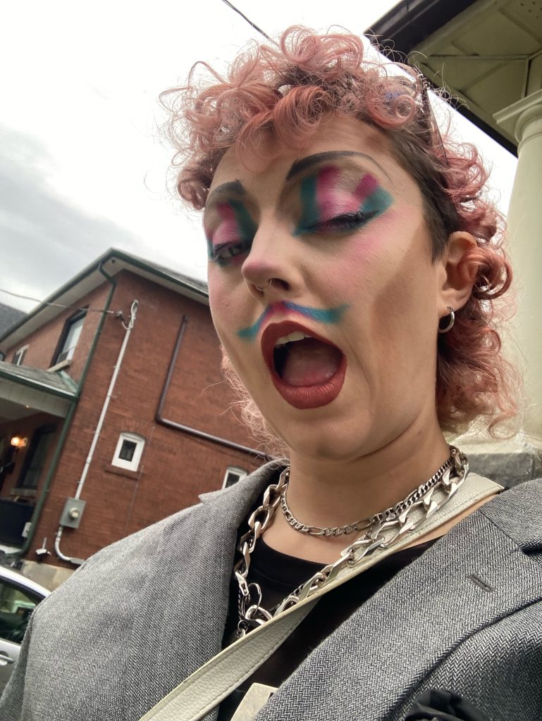 Trans person in makeup winking at the camera, urban buildings in background. 