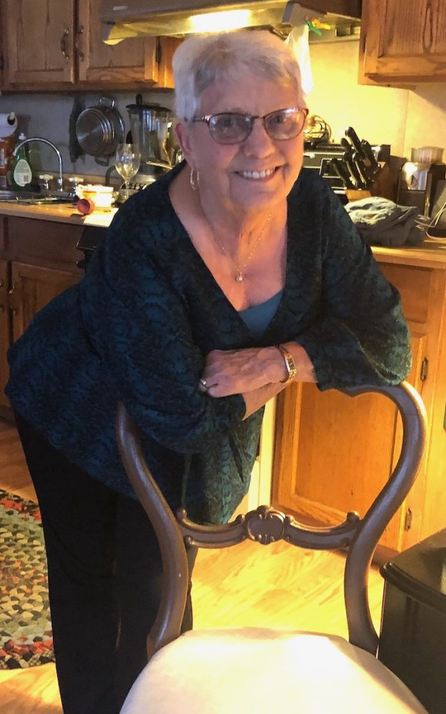 Elderly woman with grey hair and glasses leaning on a chair and smiling at the camera. 