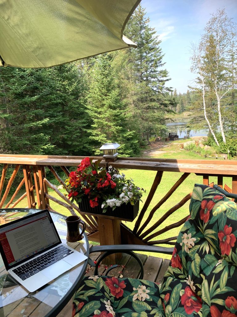 A computer on a clear table in front of a box of flowers containing begonias and pansies, a flowered chair off to the side and a green lawn and river in the foreground. 